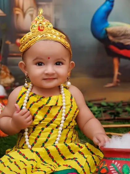 A sweet baby girl dressed as Radha, looking directly at the camera with her big, bright eyes.