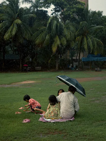 A close-up of the family having a picnic in the rain at Oval Maidan. This tighter shot focuses on the intimacy of the moment, with the father sheltering his children under the umbrella.