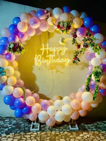 A full balloon ring in pastel colors with small floral arrangements tucked into the balloons. The 'Happy Birthday' neon sign glows in the center.