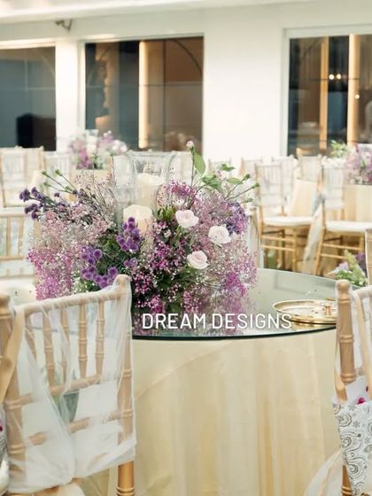 A beautiful centerpiece of purple and white flowers, including baby's breath and roses. The arrangement adds a soft pop of color to the elegantly dressed guest tables.