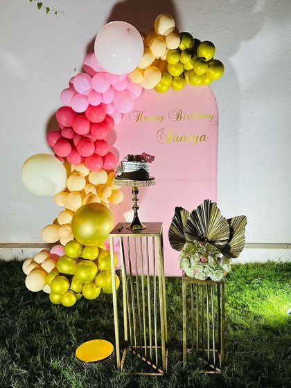 A simple yet beautiful outdoor setup on a lawn. This decoration includes a pink arched backdrop, a colorful balloon garland, and elegant golden stands for the cake and props.