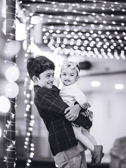 A beautiful black and white portrait of an older brother holding his smiling baby sister, with festive lights creating a lovely bokeh effect.
