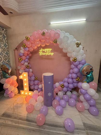 A Cocomelon-themed first birthday in a pretty pastel color scheme. The pink arch backdrop is framed with pink, purple, and white balloons, with a light-up '1' and character foils adding to the fun.
