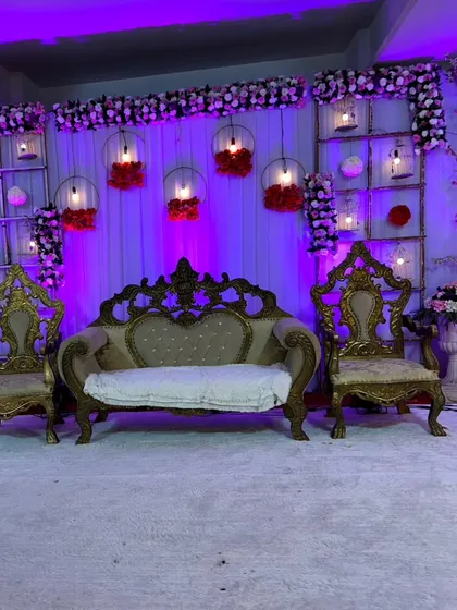 A detailed shot of the wedding reception stage, focusing on the ornate gold-trimmed sofa and chairs for the couple. The purple lighting and hanging floral decor create a romantic and luxurious atmosphere.