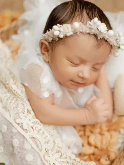 A close-up of a sleeping baby girl in a delicate white dress with a floral headband.