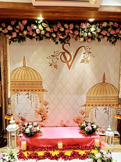 A traditional home engagement or roka setup. The backdrop features an Indian palace motif with the couple's initials, low seating, and floral decor.