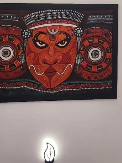 A powerful Theyyam mural hanging on a wall, illuminated by a small lamp below. This shows how the art can create a spiritual and dramatic focal point in a room.