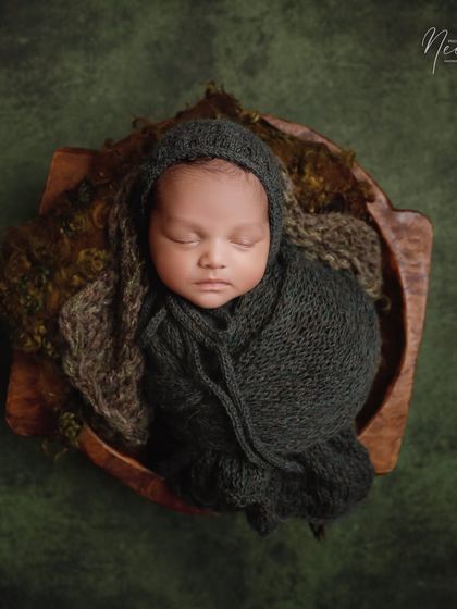 The earthy green and brown tones in this setup create a natural and calming mood. The baby is snugly wrapped in a dark grey bonnet and wrap, highlighting his peaceful face.
