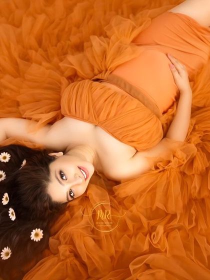 A joyful overhead portrait. Lying on a vibrant orange fabric, the mother-to-be smiles up at the camera, her hair decorated with daisies for a sweet, playful feel.