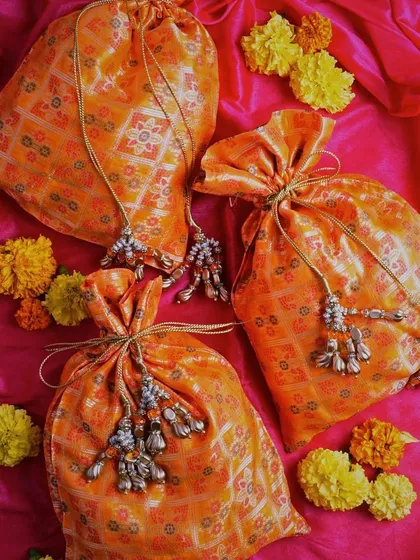A vibrant flat-lay of orange potli bags on a bright pink background. The scattered marigold flowers add to the festive and colorful mood of the shot.
