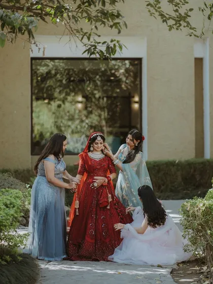 The bride, in a stunning red lehenga, gets final touch-ups from her bridesmaids in a garden setting.