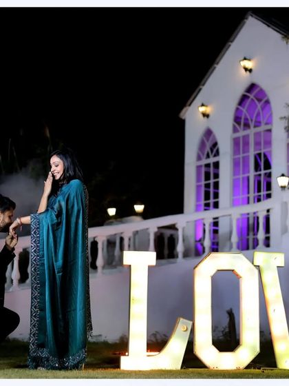 A classic proposal shot during a pre-wedding session. The groom is on one knee in front of large "LOVE" letters and a beautifully lit backdrop, creating a truly cinematic and romantic image.