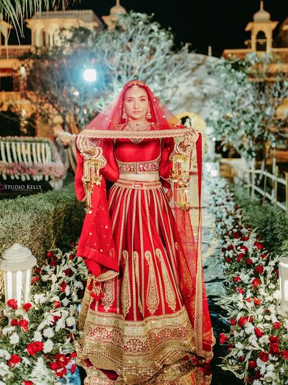 A beautiful bridal portrait of the bride in her striking red lehenga, lifting her veil under the evening lights of the palace gardens in Pushkar.