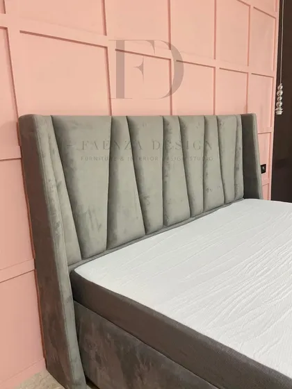 This bed, set against a pink paneled wall, features a grey velvet headboard with a striking, geometric-paneled design. The contrast of colors and shapes creates a bold, contemporary look.