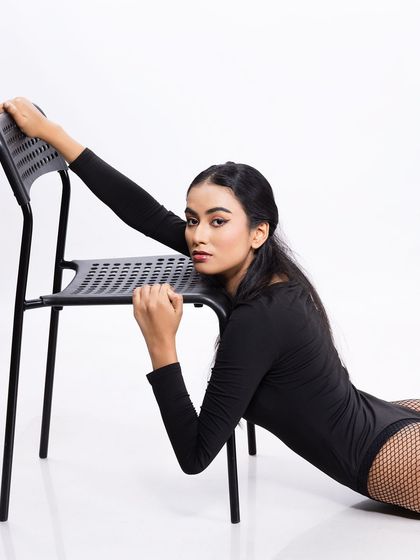 A creative fashion portrait using a simple chair as a prop. The model's pose is dynamic, showcasing the bodysuit and fishnet stockings.