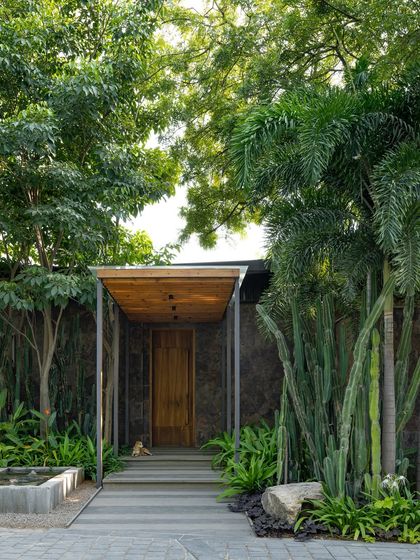 The entrance to the KEUS office, featured in Architecture+Design magazine. The image showcases the blend of modern architecture with a lush, tropical landscape, including a water feature and sculptural cacti.