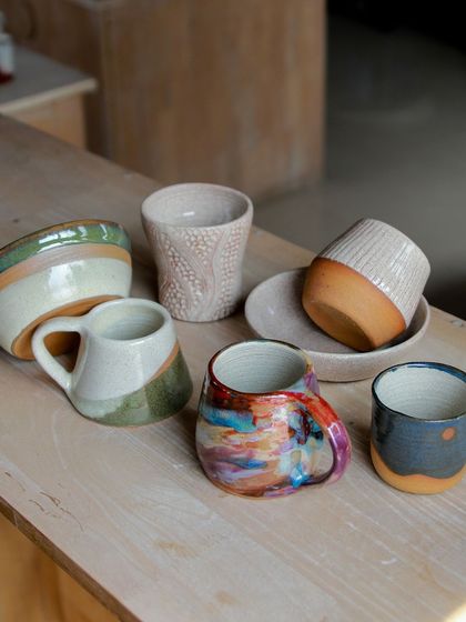 A diverse range of mugs and cups, each with a unique texture, shape, or glaze application, from a colorful abstract mug to a carved, textured cup.