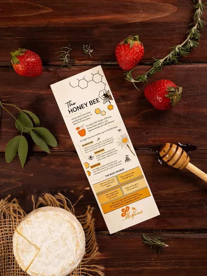 A flat lay composition for Melfons honey, featuring an informational card, fresh strawberries, and cheese. This styling suggests pairing the honey with other gourmet foods.