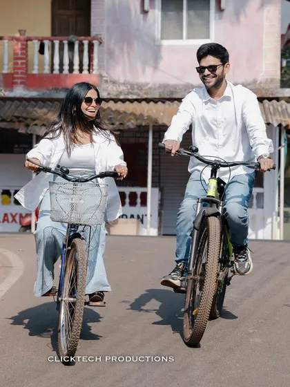 A fun and candid moment from a Goa pre-wedding shoot, showing the couple laughing as they ride bicycles down a quaint village road, capturing their natural chemistry.