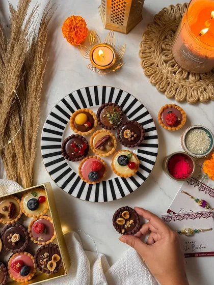 A hand selecting a mini tartlette from a platter, showcasing the variety of flavors like hazelnut chocolate, lemon, and pistachio kunafa. My mini tartlettes are an elegant and indulgent Rakhi gift.