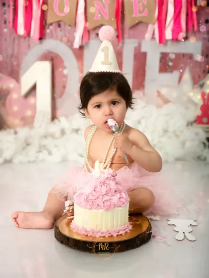 A fun and messy first birthday cake smash session. This little girl is enjoying her first taste of cake against a festive pink backdrop, a perfect way to celebrate a major milestone.