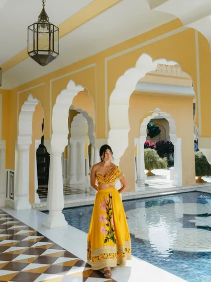 The bride stands by a palace pool, her yellow Mehendi outfit reflected in the checkered floor. The arches and architecture frame her beautifully, creating a sense of grandeur.