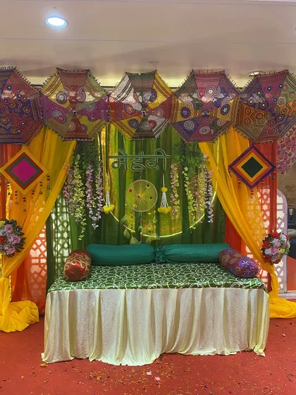 A lively mehndi setup with a green backdrop, colorful Rajasthani umbrellas, and hanging floral strings. The seating is kept simple to let the vibrant decor stand out.