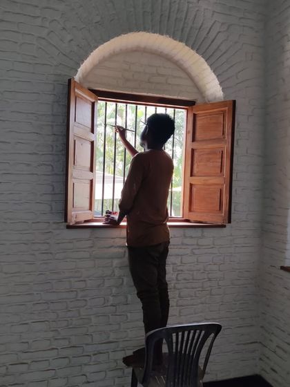 A painter applies the finishing touches to a newly installed wooden window set within a restored brick arch. The contrast between the white-painted brick and the warm wood highlights the architectural details.