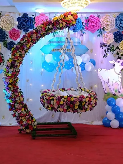 A close-up of a Janmashtami-themed cradle decoration for a naming ceremony. The cradle is adorned with flowers, set against a backdrop of paper flowers and Krishna cutouts, perfect for a traditional 'barsa'.