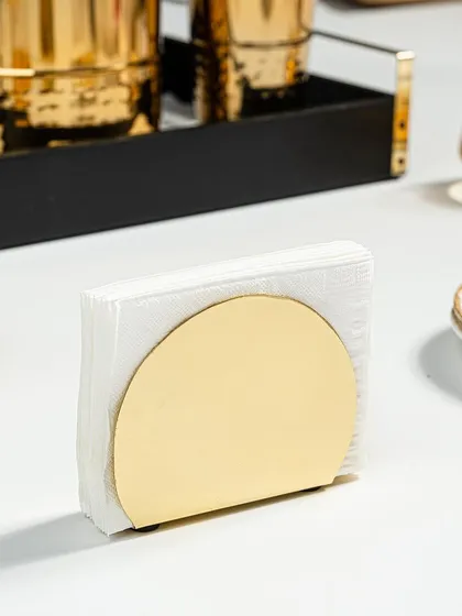 This shot showcases a semi-circular gold napkin holder as part of a larger, luxurious tablescape with gold-accented glasses and trays. It positions the product within a cohesive, high-end collection.
