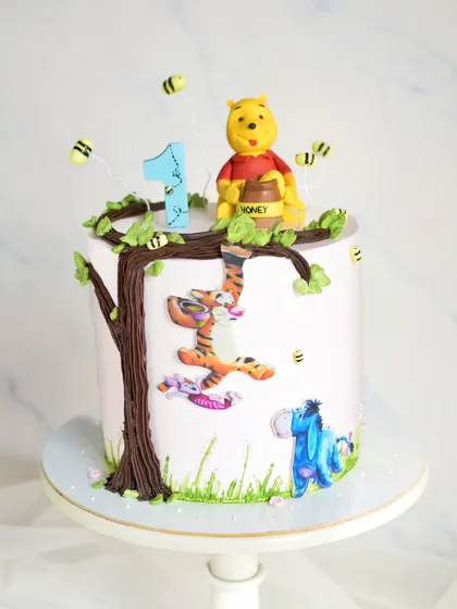 A sweet Winnie the Pooh cake for a first birthday. This design includes a hand-painted tree, edible character prints, and a cute fondant Pooh bear with his honey pot on top.