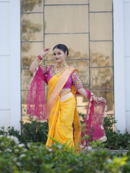A full length shot of a bride in a yellow Paithani saree with a beautiful pink shawl. The elegant pose showcases the complete look, including the intricate blouse and hairstyle.