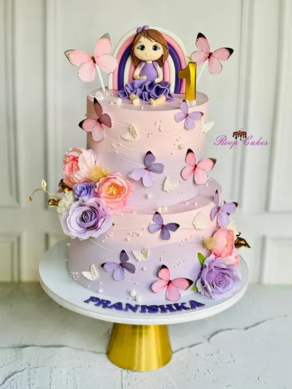 A magical two-tier cake for a very special first birthday. This design features a beautiful lavender and pink ombre effect, a custom fondant doll, and delicate butterflies, all coming together to create a fairytale celebration.