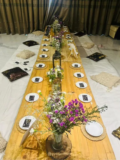 Here is a long, rustic, low seating arrangement perfect for a boho themed party. The setup uses a wooden pallet table, decorated with fresh floral centerpieces, lanterns, and place settings with macrame details, surrounded by comfortable floor cushions.
