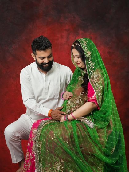 A sweet moment between the couple during their Rajasthani-themed maternity shoot. The red studio backdrop makes the vibrant colors of their outfits stand out beautifully.