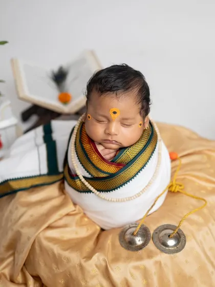 A close-up portrait focusing on the baby's face, adorned with the traditional Warkari tilak, and the textures of the wrap and cymbals.