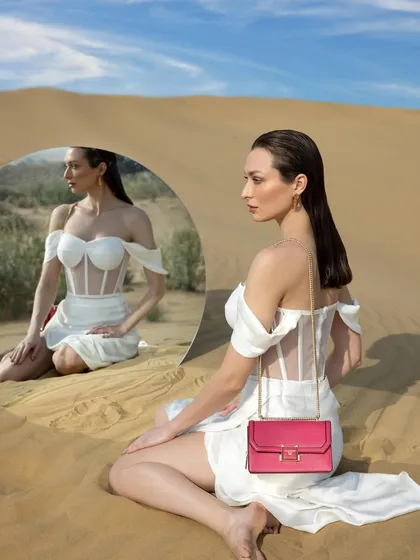 A surreal and artistic campaign shot for a luxury handbag brand. The model in a white corset dress sits in the desert, her reflection captured in a mirror, with a pop of color from the pink bag.