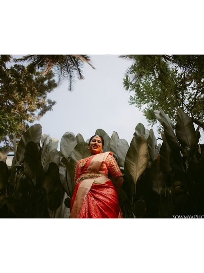 A low-angle shot of a bride in a red saree, standing amidst large green leaves. The perspective is unique and striking.