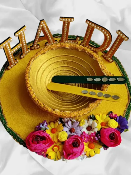 This Haldi platter features a beautiful 'HALDI' sign with pearl detailing, a golden bowl, and a base decorated with a burst of colourful flowers.
