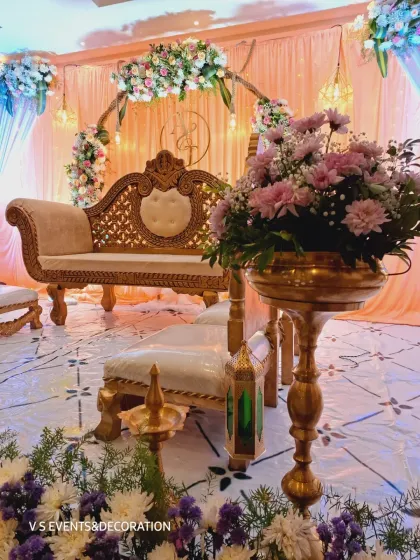 This engagement setup combines traditional and modern elements. The stage features a classic diwan-style sofa, traditional brass lamps (diya), and Moroccan-style lanterns, all set against a backdrop of soft peach and blue drapes with a floral arch.