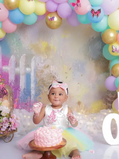 A baby girl, dressed as a unicorn, gets her first taste of her birthday cake. This cake smash photo is full of color and excitement, set against a whimsical, pastel-colored backdrop.
