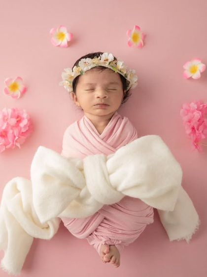 A full-body shot of a newborn wrapped in pink with a large white bow, lying on a pink background scattered with flower petals.