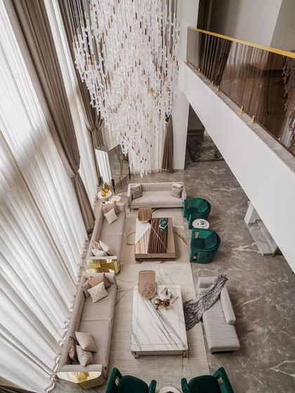 An aerial view of the double-height living room, showcasing the grand scale and thoughtful furniture layout. The long sofas and emerald armchairs are arranged to facilitate conversation, all under the glow of a massive crystal chandelier.