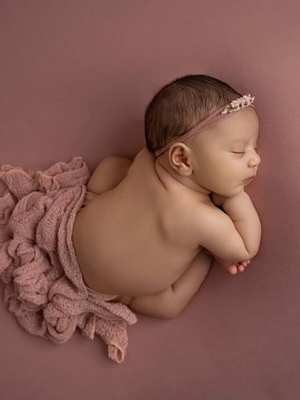 An overhead view of a baby girl in the 'taco' pose, with a simple pink wrap and headband against a matching mauve background.