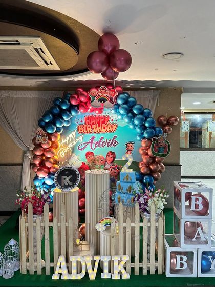 A Cocomelon party with a circular backdrop and a metallic balloon arch. The setup includes themed plinths, 'BABY' blocks, and a personalized name sign.