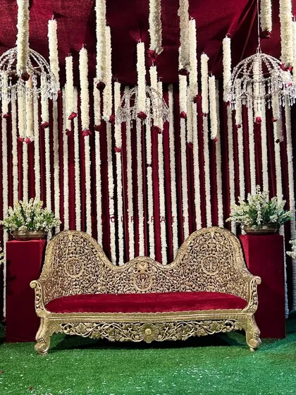 A full stage view of the traditional Godh Bharai ceremony. The ornate gold sofa sits on a green lawn against a backdrop of red velvet and hanging white floral garlands with crystal chandeliers.