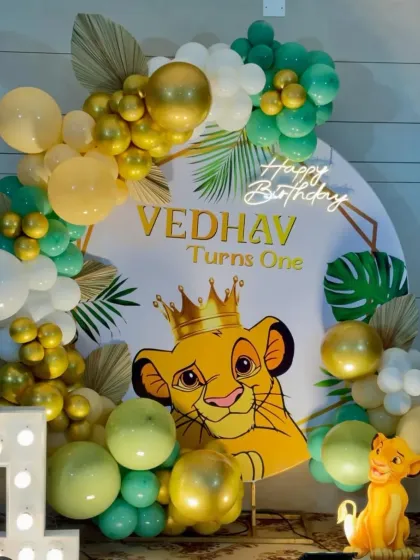 A cute Simba-themed first birthday with a character backdrop, a "1" marquee number, and a balloon arch in green, yellow, and gold.