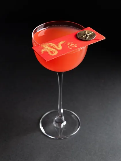 An elegant red cocktail in a coupe glass, garnished with a decorative red envelope for a themed event. The clean, black background makes the colour of the drink the absolute hero.