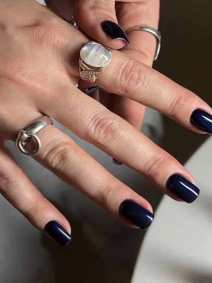 A close-up of a chic, dark manicure. The deep navy polish on these short nails, paired with silver rings, creates a stylish and modern aesthetic.