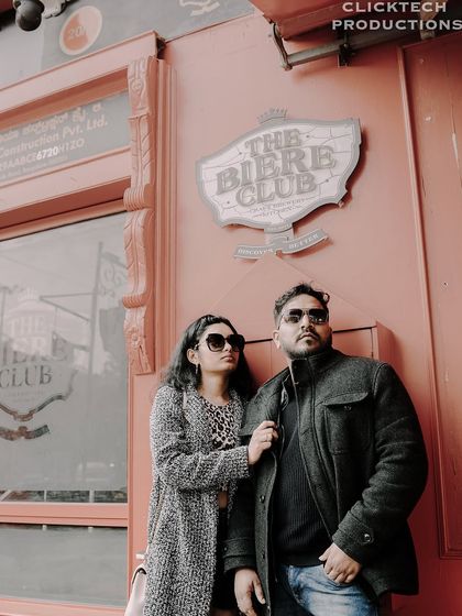 A low-angle shot of a couple in stylish coats, posing against the wall of 'The Biere Club', creating a cool, urban portrait.